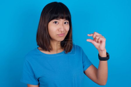 Foto de Mujer asiática joven disgustado usando camiseta azul contra fondo azul formas poco signo de mano demuestra algo no muy grande. Concepto de lenguaje corporal. - Imagen libre de derechos