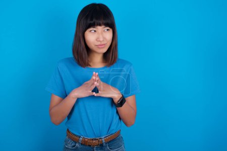 Photo for Young asian woman wearing blue t-shirt against blue background steepls fingers and looks mysterious aside has great evil plan in mind - Royalty Free Image