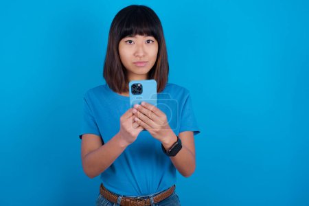 Téléchargez les photos : Portrait de sérieux confiant jeune asiatique femme portant bleu t-shirt sur fond bleu tenant téléphone dans les deux mains - en image libre de droit