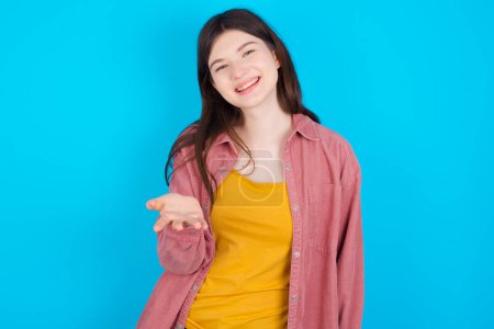 Foto de Joven chica caucásica con camisa rosa aislada sobre fondo azul sonriendo amigable ofreciendo algo con la mano abierta o apretón de manos como saludo y bienvenida. Negocios exitosos. - Imagen libre de derechos
