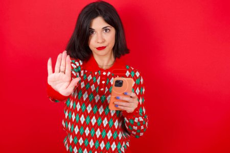 Photo for Brunette caucasian woman wearing christmas sweater over red background using and texting with smartphone with open hand doing stop sign with serious and confident expression, defense gesture - Royalty Free Image
