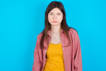 Photo for Gloomy, bored young caucasian girl wearing pink shirt isolated over blue background frowns face looking up, being upset with so much talking hands down, feels tired and wants to leave. - Royalty Free Image