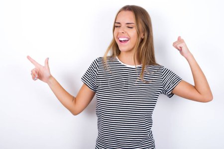 Foto de Alegre hermosa chica rubia con camiseta a rayas sobre fondo blanco mostrando espacio de copia anuncio celebrando suerte - Imagen libre de derechos