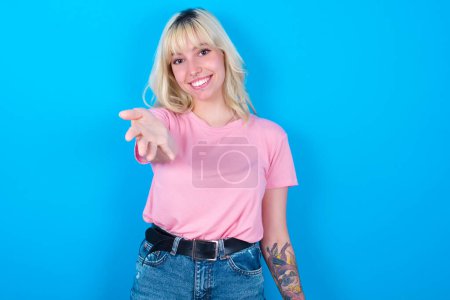 Photo for Caucasian girl wearing pink t-shirt isolated over blue background smiling friendly offering something with open hand or handshake as greeting and welcoming. Successful business. - Royalty Free Image