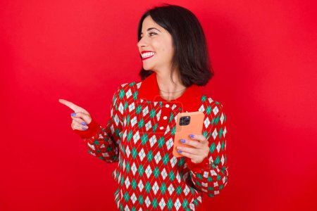Téléchargez les photos : Ouah ! excitée femme caucasienne brune portant chandail de Noël sur fond rouge montrant téléphone mobile avec geste de la main ouverte - en image libre de droit