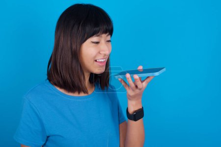 Téléchargez les photos : Souriante jeune femme asiatique portant un t-shirt bleu sur fond bleu envoyant un message vocal sur son téléphone intelligent. Concept de communication et nouvelles technologies. - en image libre de droit
