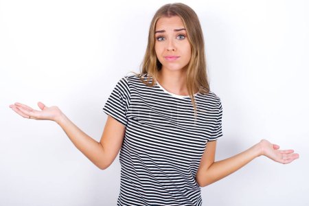 Photo for Careless attractive beautiful blonde girl wearing striped t-shirt on white background shrugging shoulders, oops. - Royalty Free Image