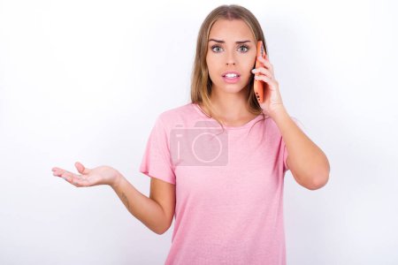 Téléchargez les photos : Jeune fille caucasienne portant T-shirt rose sur fond blanc parlant au téléphone stressé avec la main sur le visage, choqué par la honte et le visage surprise, en colère et frustré. Peur et contrariété par erreur. - en image libre de droit