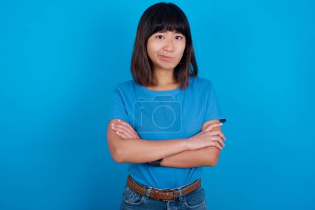 Téléchargez les photos : Photo d'une jeune femme asiatique en colère portant un t-shirt bleu sur fond bleu croisant les bras. Regardant la caméra avec une expression déçue. - en image libre de droit