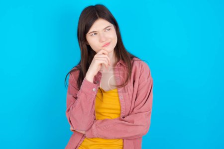 Photo for Young caucasian girl wearing pink shirt isolated over blue background with hand under chin and looking sideways with doubtful and skeptical expression, suspect and doubt. - Royalty Free Image