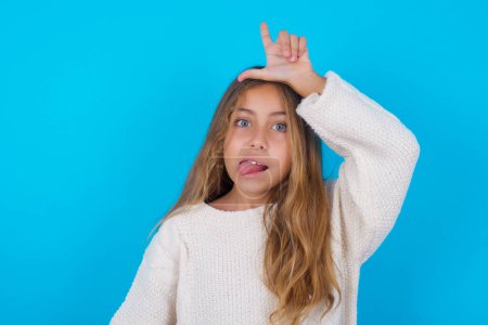Photo for Pretty teen girl gestures with finger on forehead makes loser gesture makes fun of people shows tongue - Royalty Free Image