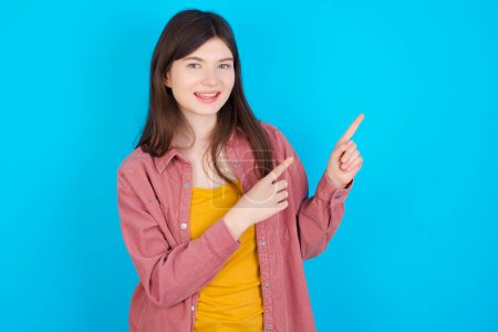 Photo for Young caucasian girl wearing pink shirt isolated over blue background with positive expression, indicates with fore finger at blank copy space for your promotional text or advertisement. - Royalty Free Image