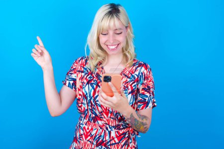 Téléchargez les photos : Sourire fille caucasienne portant robe florale isolé sur fond bleu pointant doigt à l'espace vide tenant téléphone dans une main - en image libre de droit