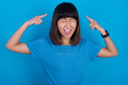 Photo for Photo of crazy young asian woman wearing blue t-shirt against blue background screaming and pointing with fingers at hair closed eyes - Royalty Free Image