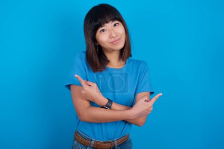 Foto de Joven mujer asiática vistiendo camiseta azul contra fondo azul cruza brazos y puntos en diferentes lados duda entre dos artículos o variantes. Necesita ayuda con la decisión - Imagen libre de derechos