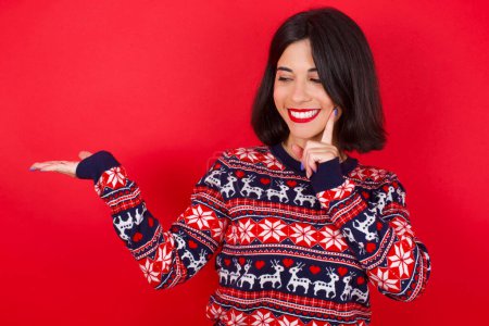 Téléchargez les photos : Drôle de femme caucasienne brune portant un pull de Noël sur fond rouge tenant la paume ouverte nouveau produit. Je veux l'acheter.! - en image libre de droit