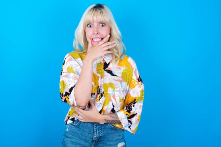 Foto de Chica caucásica vistiendo camisa floral aislada sobre fondo azul cubriendo la boca con las manos asustadas de algo o alguien mordiendo uñas - Imagen libre de derechos