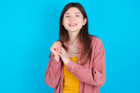 Photo for Dreamy charming young caucasian girl wearing pink shirt isolated over blue background with pleasant expression, keeps hands crossed near face, excited about something pleasant. - Royalty Free Image