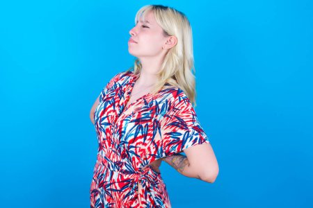 Photo for Caucasian girl wearing floral dress isolated over blue background got back pain - Royalty Free Image