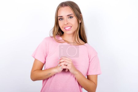 Photo for Happy Young Caucasian girl wearing pink T-shirt on white background stands against orange studio wall keeps hands on heart, swears be loyal, expresses gratitude. Honesty concept. - Royalty Free Image