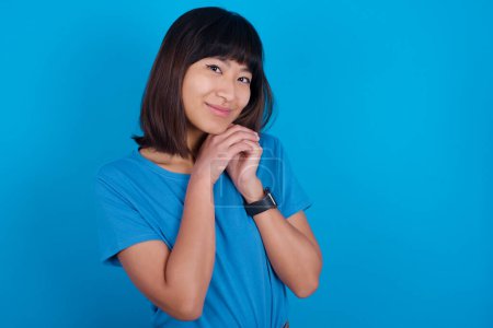 Foto de Encantadora joven asiática seria que usa camiseta azul contra fondo azul mantiene las manos cerca de la cara sonríe tiernamente a la cámara - Imagen libre de derechos