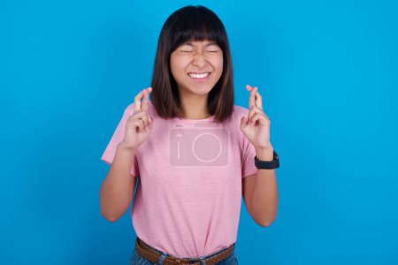 Téléchargez les photos : Joyeuse jeune femme asiatique portant un t-shirt rose sur fond bleu serre les dents, lève les doigts croisés, fait un souhait souhaitable, attend de bonnes nouvelles, je dois gagner. - en image libre de droit