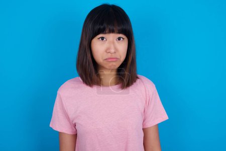 Foto de Joven mujer asiática vistiendo camiseta rosa sobre fondo azul llorando desesperado y deprimido con lágrimas en los ojos sufriendo dolor y depresión. Triste expresión facial y concepto de emoción. - Imagen libre de derechos