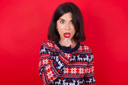 Foto de Nervioso desconcertado morena caucásica mujer usando navidad suéter sobre fondo rojo abre la boca de sorpresa, reacciona en noticias repentinas. - Imagen libre de derechos