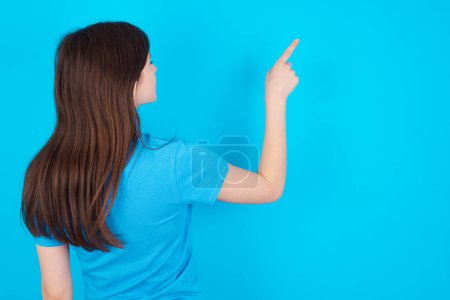 Foto de Joven chica caucásica vistiendo camiseta azul aislado sobre fondo azul estudio apuntando a objeto en el espacio de copia, vista trasera. Da la espalda. - Imagen libre de derechos