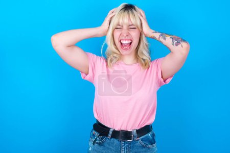 Photo for Shocked panic caucasian girl wearing pink t-shirt isolated over blue background holding hands on head and screaming in despair and frustration. - Royalty Free Image
