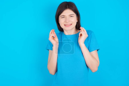 Téléchargez les photos : Jeune fille caucasienne portant bleu t-shirt isolé sur fond bleu studio tenant les doigts croisés avec expression inquiète espérant patron n'a pas remarqué d'erreurs au travail. - en image libre de droit