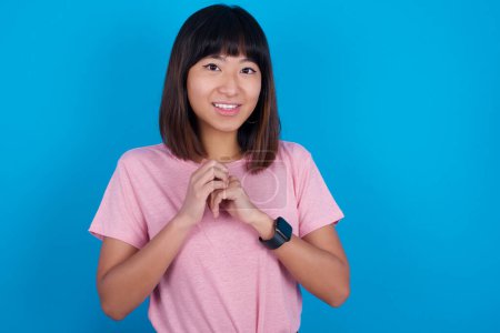 Téléchargez les photos : Rêveuse charmante jeune femme asiatique portant un t-shirt rose sur fond bleu avec une expression agréable, garde les mains croisées près du visage, excitée par quelque chose d'agréable. - en image libre de droit