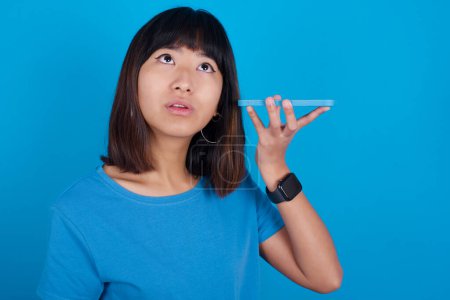 Photo for Smiling young asian woman wearing blue t-shirt against blue background listening a voice message from her smartphone. Communication and technology concept. - Royalty Free Image