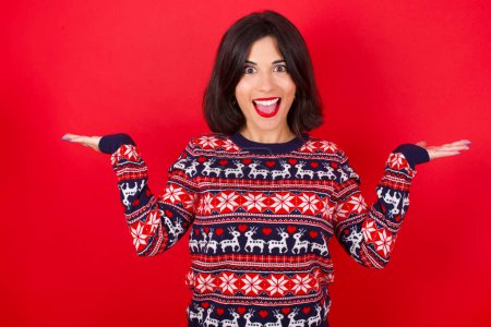 Téléchargez les photos : Femme caucasienne brune portant un pull de Noël sur fond rouge célébrant fou et étonné pour le succès avec les bras levés et les yeux ouverts hurlant excités. Concept gagnant - en image libre de droit