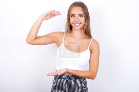 Young Caucasian girl wearing white tank top on white background gesturing with hands showing big and large size sign, measure symbol.
