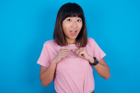 Photo for Portrait of desperate and shocked young asian woman wearing pink t-shirt against blue background looking panic, holding hands near face, with mouth wide open. - Royalty Free Image