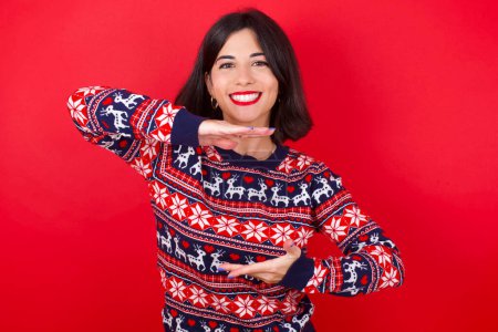 Téléchargez les photos : Brune femme caucasienne portant chandail de Noël sur fond rouge geste avec les mains montrant grand et grand signe de taille, symbole de mesure. Souriant en regardant la caméra. - en image libre de droit