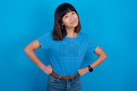 Photo for Funny frustrated young asian woman wearing blue t-shirt against blue background holding hands on waist and silly looking at awkward situation. - Royalty Free Image