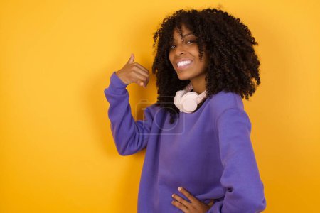 Téléchargez les photos : Portrait de jeune femme afro-américaine expressive avec écouteurs sur fond jaune montrant appel moi geste - en image libre de droit