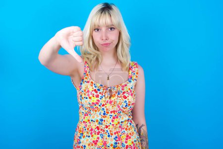 Photo for Caucasian girl wearing floral dress isolated over blue background looking unhappy and angry showing rejection and negative with thumbs down gesture. Bad expression. - Royalty Free Image