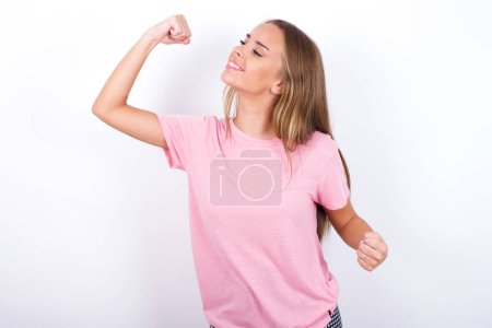 Téléchargez les photos : Attrayant jeune fille caucasienne portant T-shirt rose sur fond blanc célébrant une victoire frapper l'air avec ses poings et un sourire de dents rayonnantes - en image libre de droit