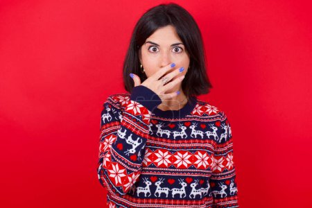 Téléchargez les photos : Femme caucasienne brune émotionnelle portant un pull de Noël sur fond rouge halète d'étonnement, couvre la bouche ouverte avec la paume, semble choqué à la caméra. Concept de réaction - en image libre de droit