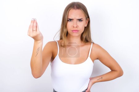 Foto de What the hell are you talking about. Shot of frustrated Young Caucasian girl wearing white tank top on white background gesturing with raised hand doing Italian gesture, frowning, being displeased and confused with dumb question. - Imagen libre de derechos