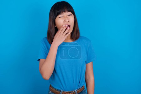 Foto de Mujer asiática joven y somnolienta con camiseta azul sobre fondo azul bostezando con el pelo desordenado, sintiéndose cansada después de la noche sin dormir, bostezando, cubriendo la boca con palma. - Imagen libre de derechos
