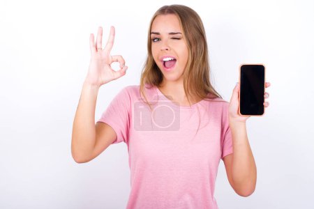 Photo for Excited Young Caucasian girl wearing pink T-shirt on white background showing smartphone blank screen, blinking eye and doing ok sign with hand.  Advertisement concept. - Royalty Free Image