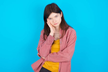 Photo for Very bored young caucasian girl wearing pink shirt isolated over blue background holding hand on cheek while support it with another crossed hand, looking tired and sick. - Royalty Free Image