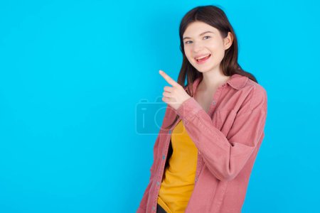 Téléchargez les photos : Jeune fille caucasienne portant chemise rose isolé sur fond bleu pointant loin et vous souriant. Regarde là-bas.! - en image libre de droit