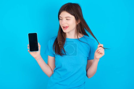 Photo for Photo of nice pretty young caucasian girl wearing blue t-shirt isolated over blue studio background demonstrate phone screen hold hair tails - Royalty Free Image