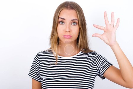beautiful blonde girl wearing striped t-shirt on white background purses lip and gestures with hand, shows something very little.