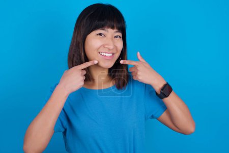 Téléchargez les photos : Dents blanches droites saines fortes. Gros plan portrait de heureuse jeune femme asiatique portant un t-shirt bleu sur fond bleu avec un sourire rayonnant pointant sur des dents blanches claires parfaites. - en image libre de droit